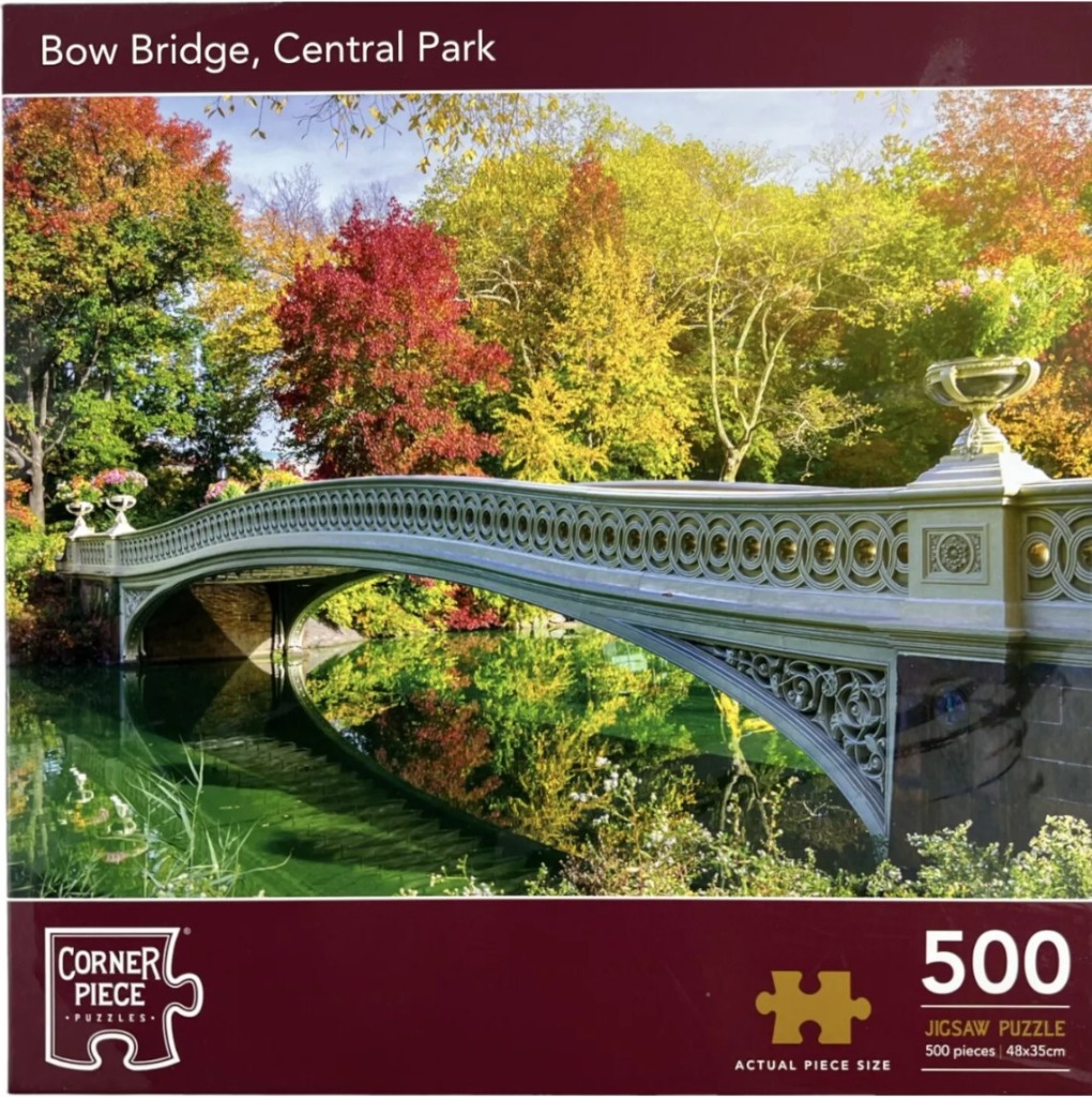 Photograph of the Bow Bridge in Central Park surrounded by colorful autumn trees reflected in the calm water.