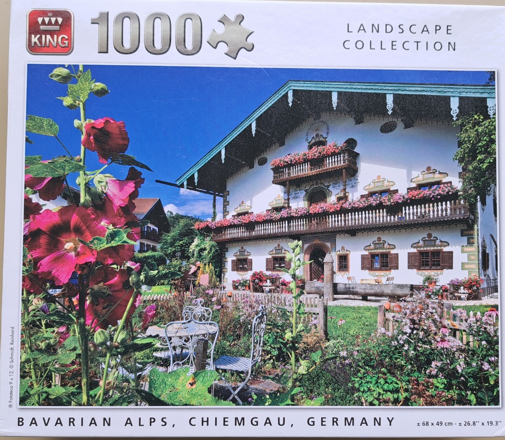 A traditional Bavarian farmhouse with flower-filled balconies, a garden with hollyhocks, and a bright blue sky.