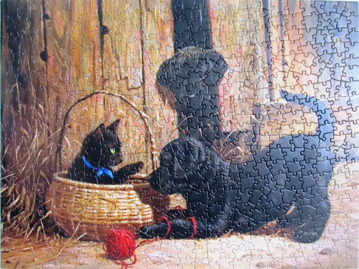 A black kitten in a wicker basket and a black puppy playing on a barn floor.