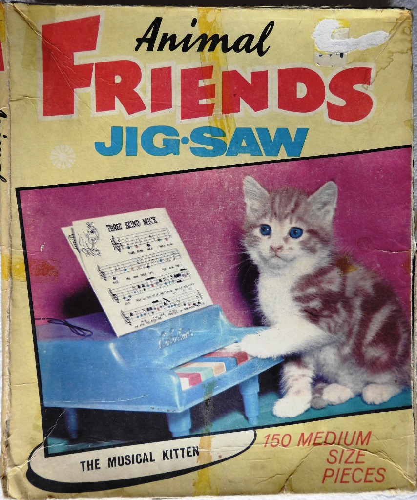 A fluffy grey and white kitten sitting next to a small blue toy piano with sheet music for Three Blind Mice.