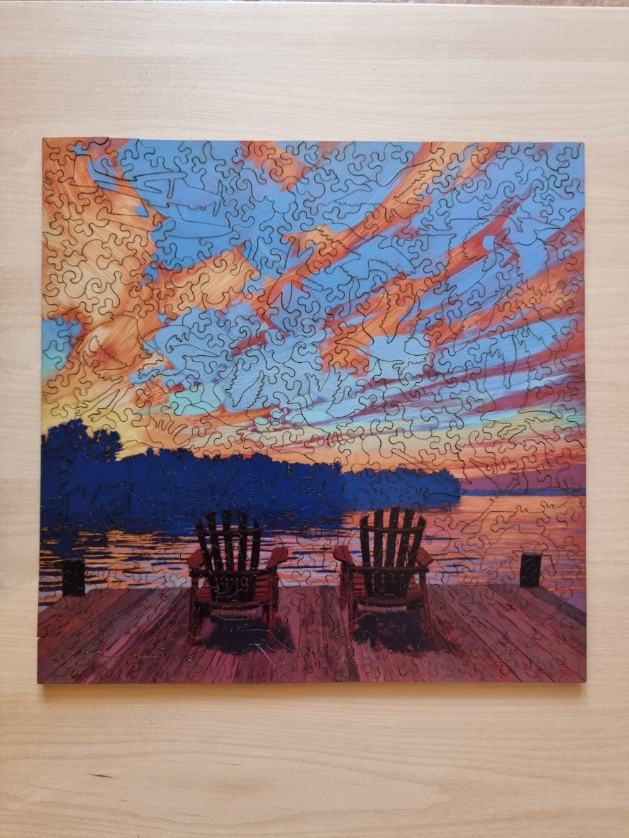 Two Adirondack chairs on a wooden dock overlooking a lake at sunset.