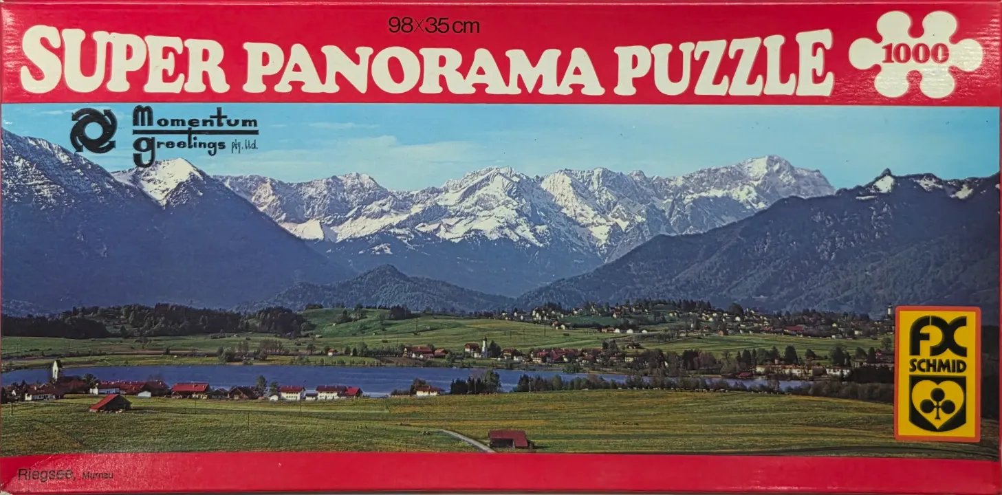 Panoramic landscape of Riegsee, Bavaria, featuring a lake and village before snow-capped mountains.