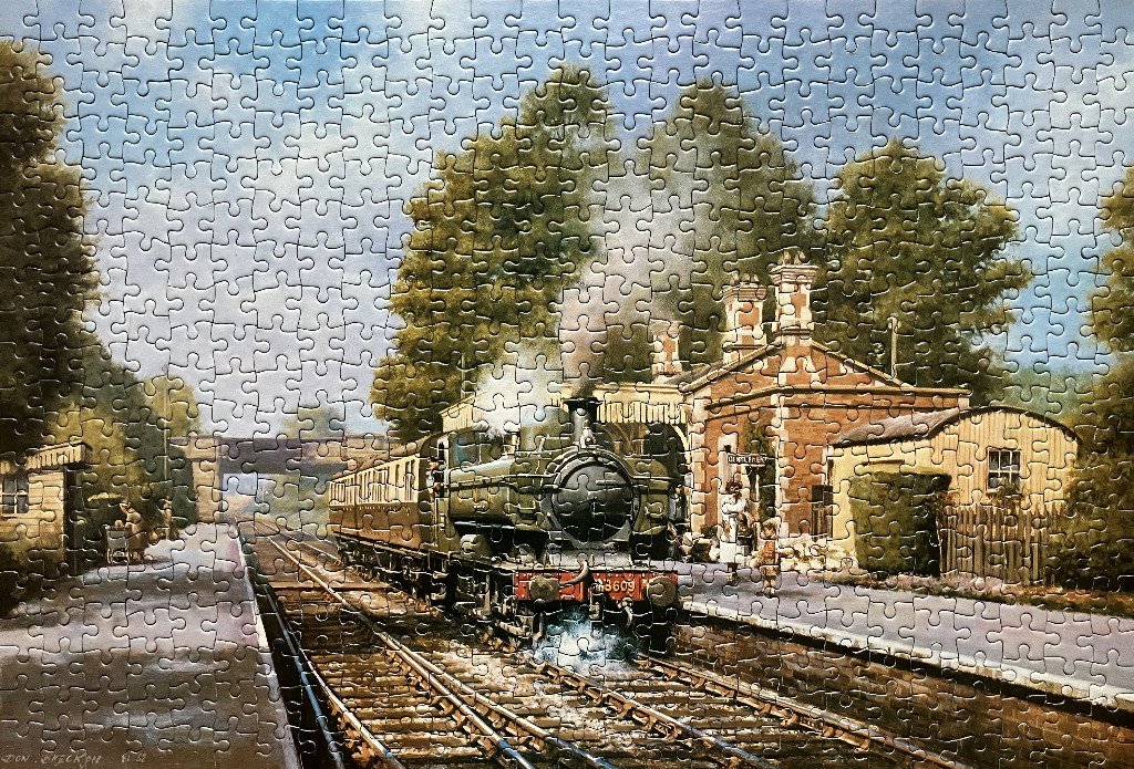 A vintage steam locomotive pulling into a brick-built country railway station on a sunny day.