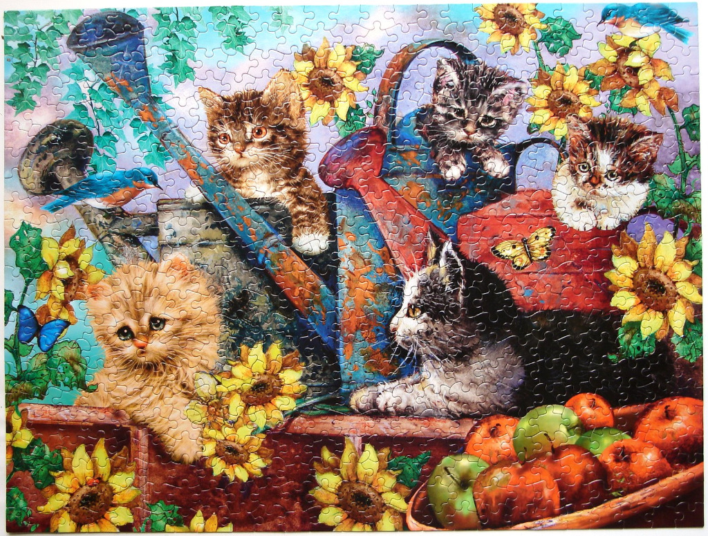 Four kittens nestled amongst sunflowers, watering cans, and a bowl of apples in a garden setting.