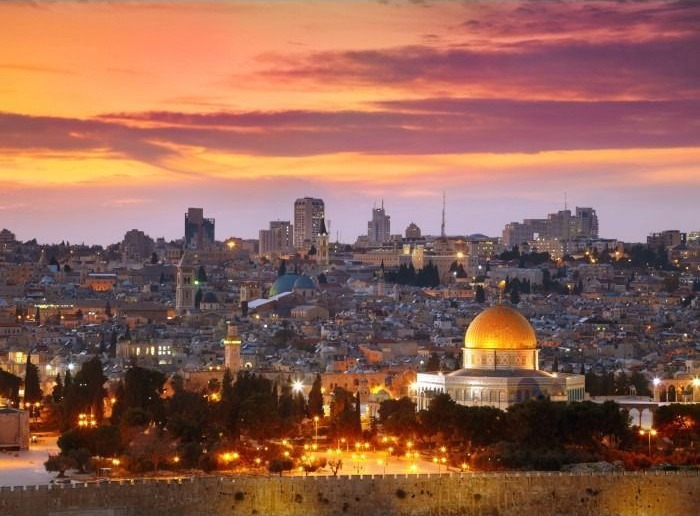 The golden Dome of the Rock in Jerusalem at sunset, surrounded by the city skyline under a colorful orange sky.