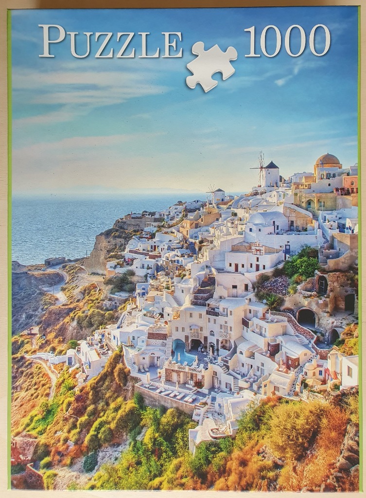A sunny photograph of white buildings clinging to a rocky Greek cliffside overlooking the blue sea.