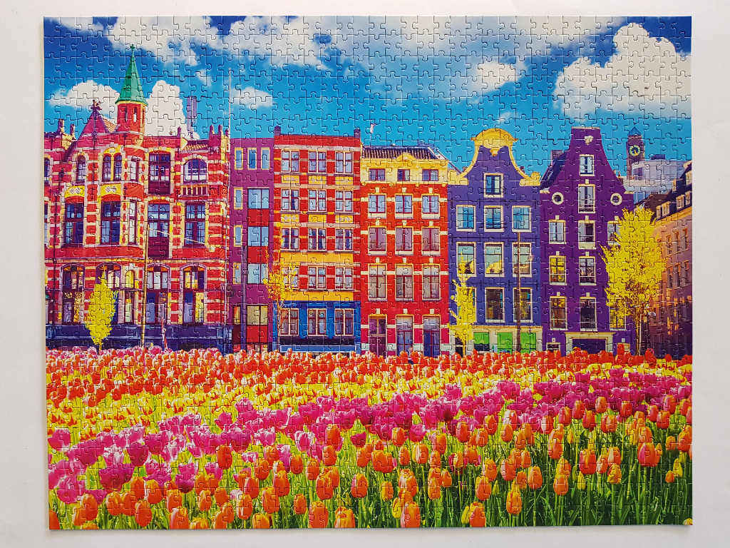 A row of colorful Dutch canal houses behind a vibrant field of pink, orange, and yellow tulips under a blue sky.