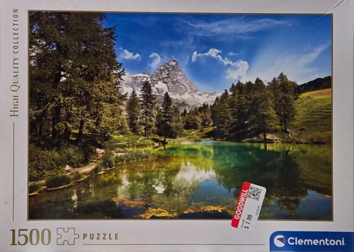 A clear mountain lake reflecting a towering, snow-capped rocky peak framed by evergreen trees under a bright blue sky.