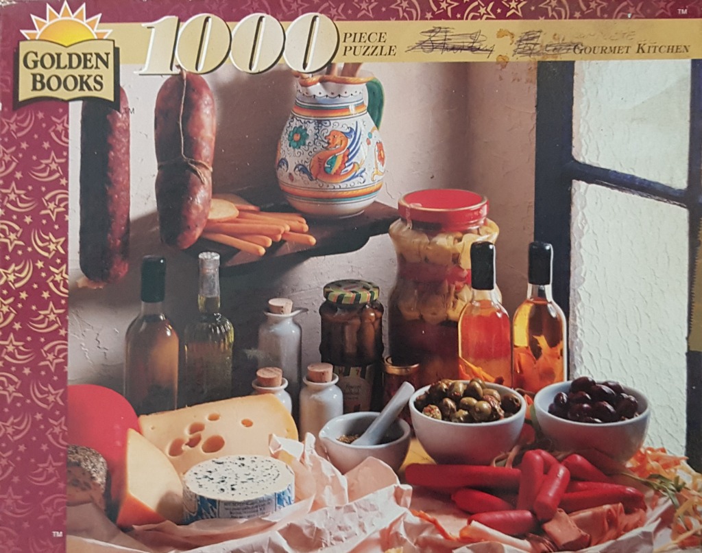A still life photograph of gourmet kitchen ingredients including various cheeses, sausages, olives, and bottled preserves.