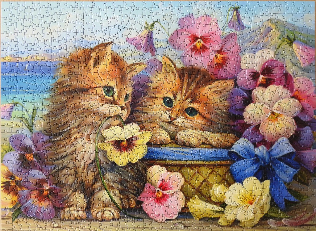 Two fluffy tabby kittens sitting in a basket surrounded by colorful pansies and a blue bow.