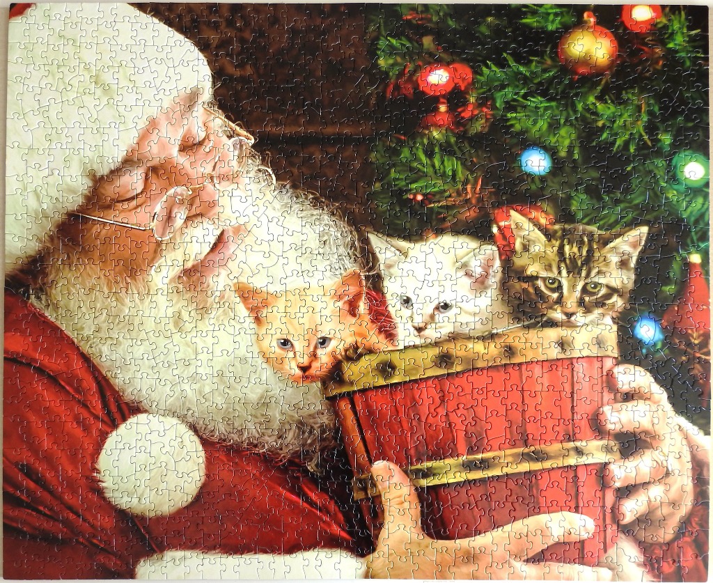 Santa Claus holding a wooden crate containing three kittens in front of a decorated Christmas tree.