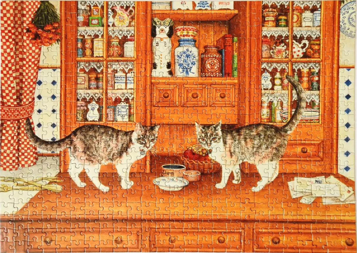 Two cats stand on a kitchen counter in front of a rustic wooden hutch filled with jars and decorations.