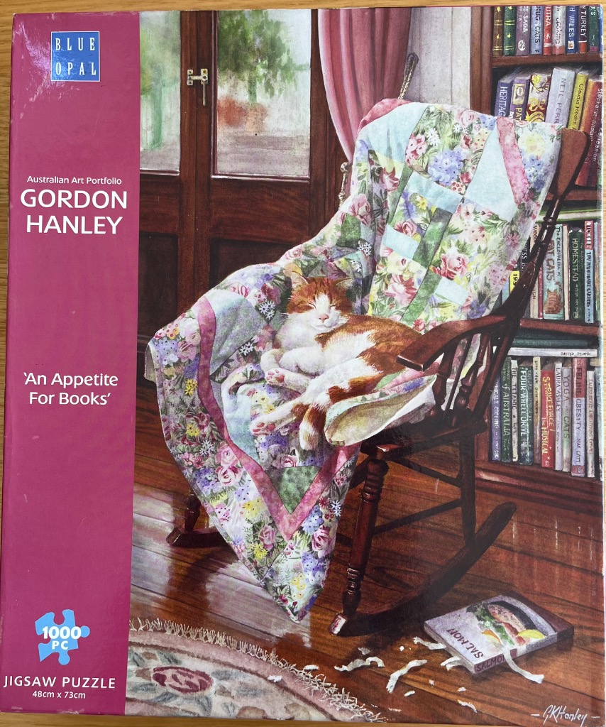 A ginger and white cat sleeping on a floral quilt in a wooden rocking chair next to a bookshelf.