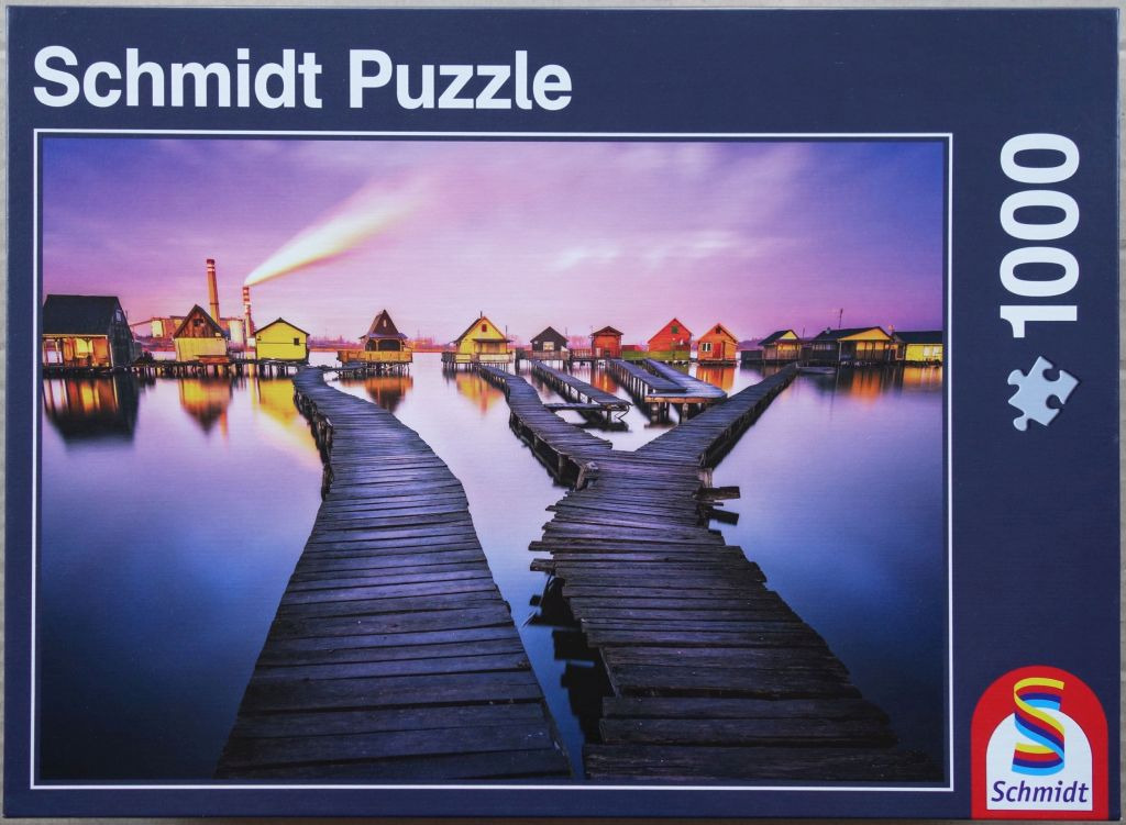 A row of colorful small wooden cabins on stilts over water with a bifurcating wooden walkway at sunset.