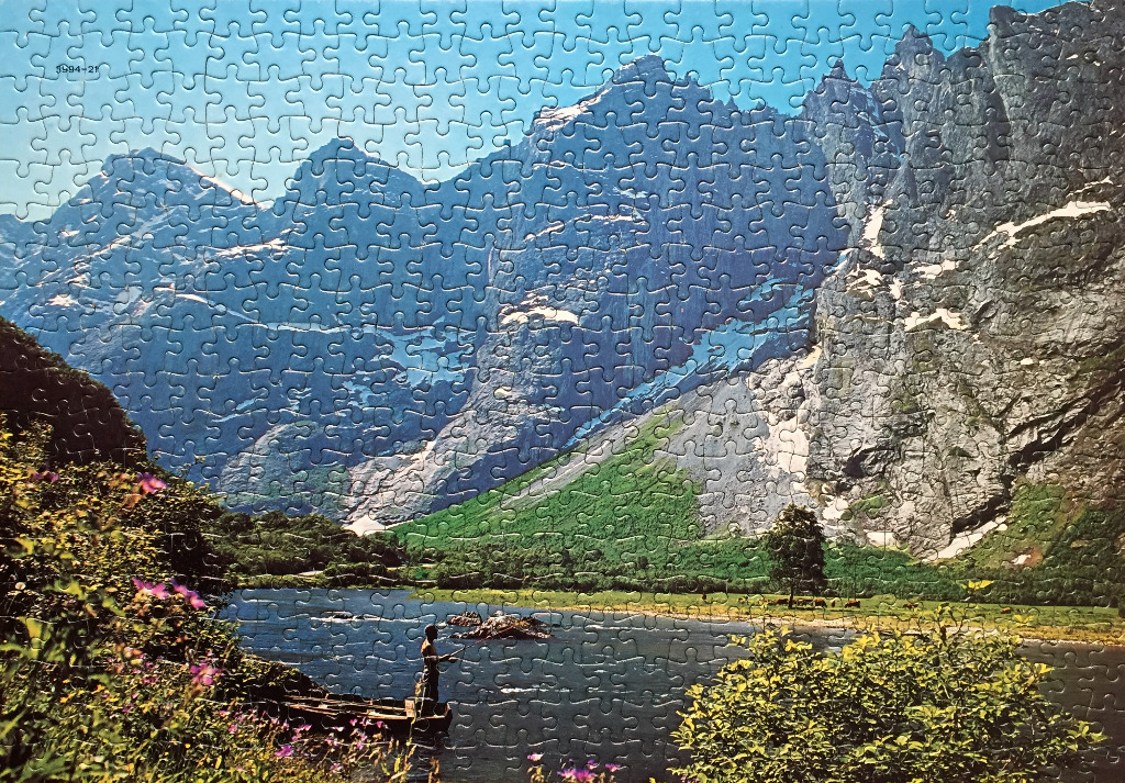 A lone fisherman in a small boat on a river surrounded by towering, rugged mountains under a clear blue sky.