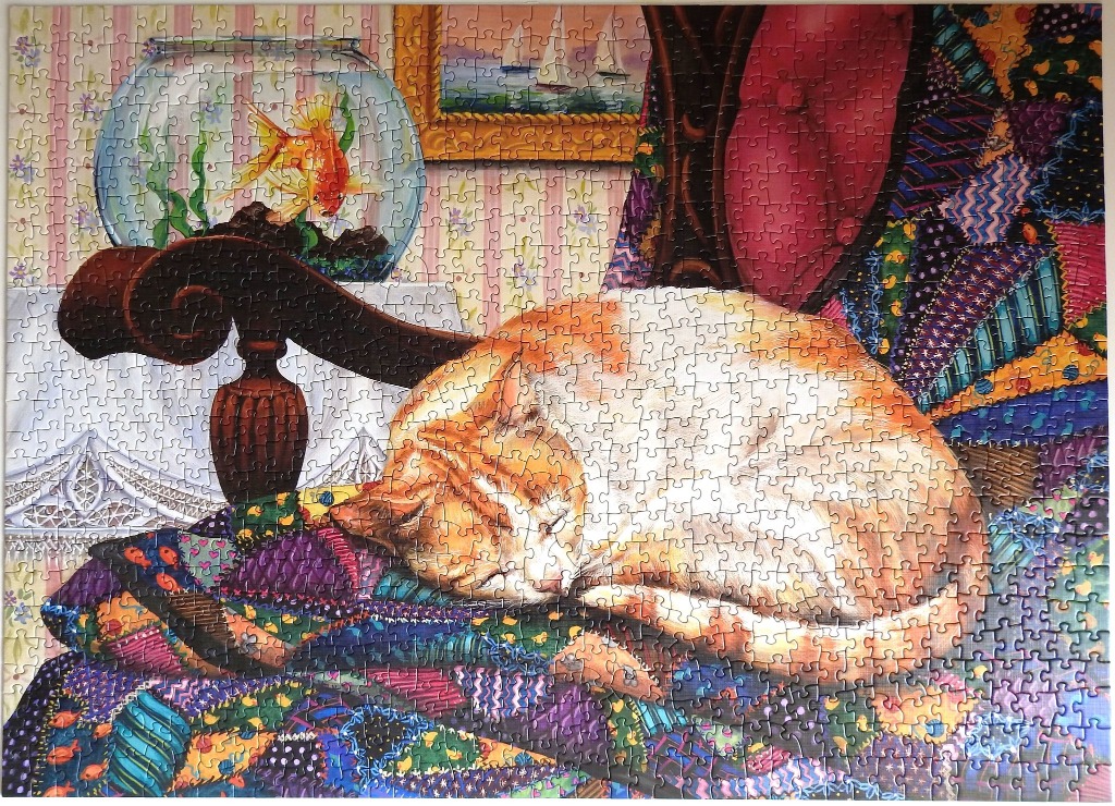 An orange tabby cat sleeping on a colorful patchwork quilt near a fishbowl in a room with striped wallpaper.