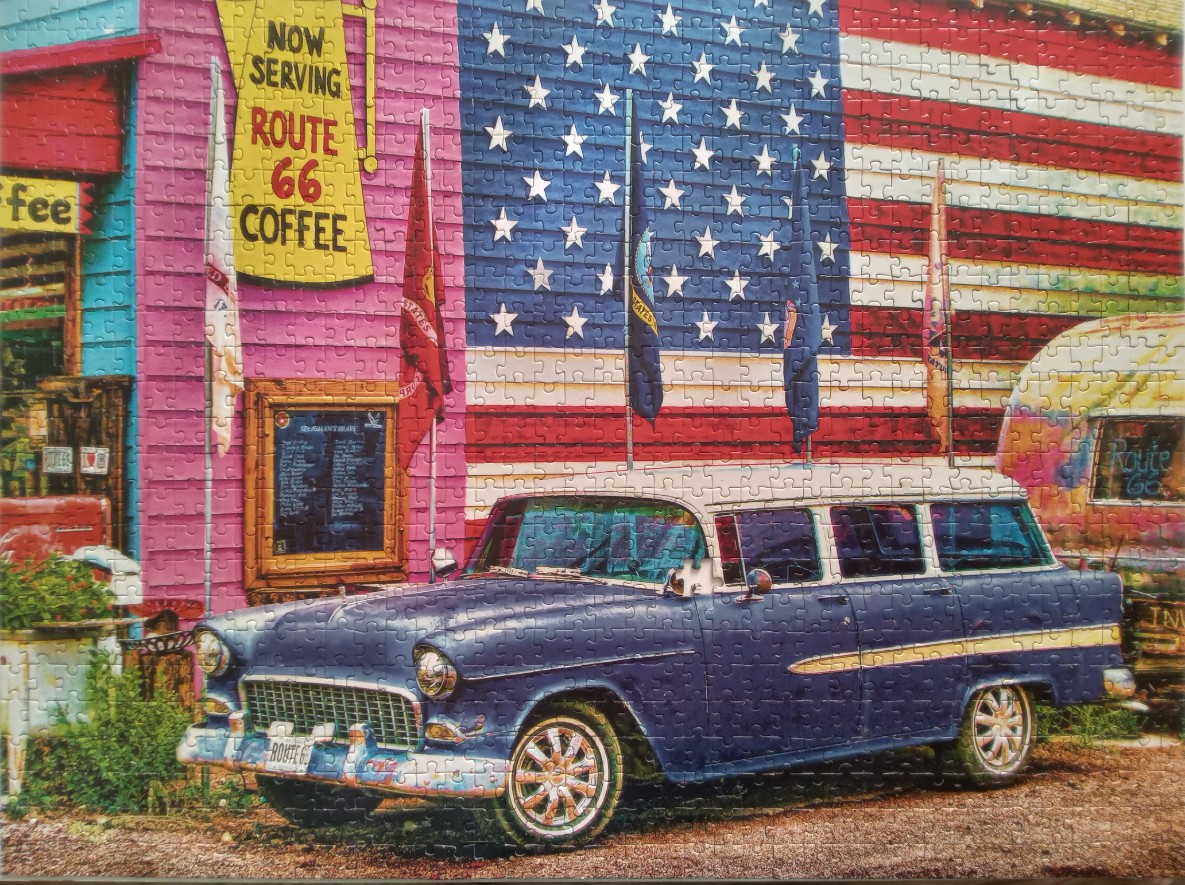 A vintage blue station wagon parked in front of a colorful Route 66 roadside diner and a large American flag.