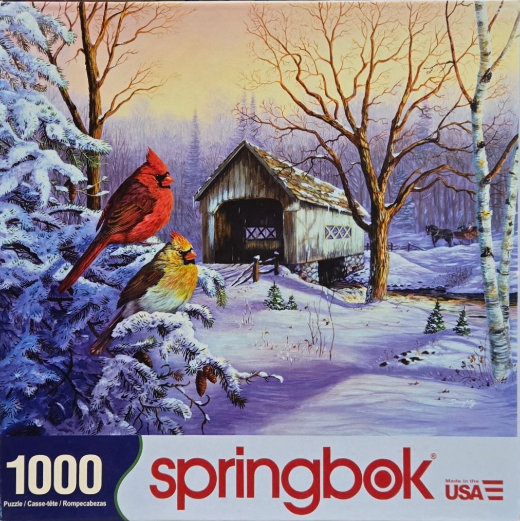 Two northern cardinals perched on a snowy pine branch with a covered bridge in the background.