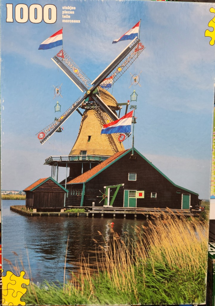 A traditional Dutch windmill with a thatched roof and brown wooden building sits beside a canal on a sunny day.