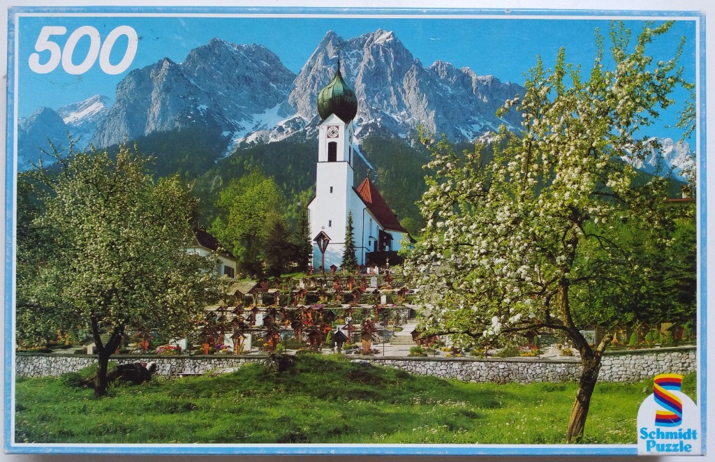 A white church with a green onion dome sits in front of jagged, snow-dusted mountains in an Alpine spring scene.