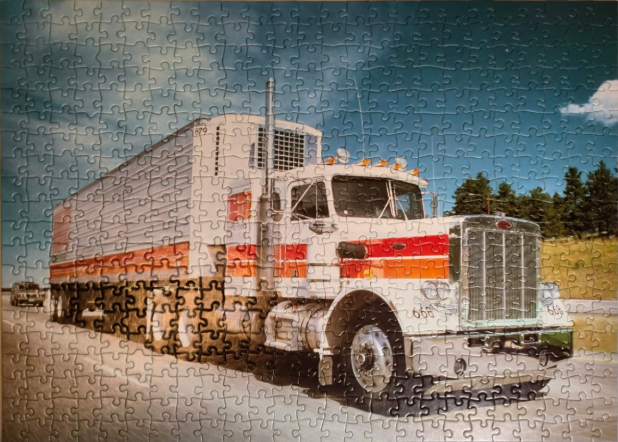 A vintage semi-truck with a white and orange trailer driving on a highway under a cloudy blue sky.