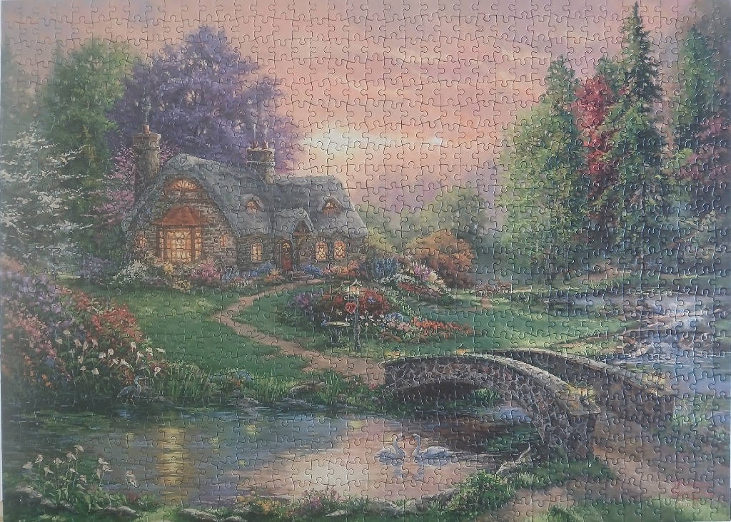 A cozy stone cottage with a thatched roof sits beside a tranquil pond and stone bridge at sunset.