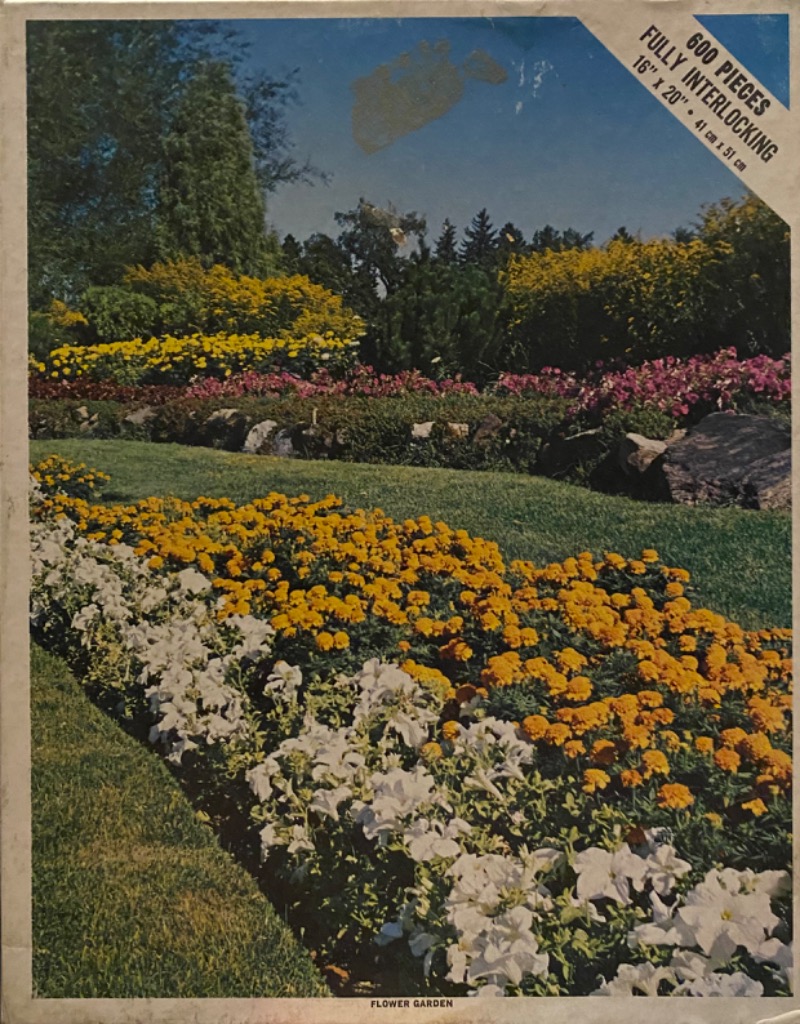 A lush summer garden scene with beds of white and orange flowers leading to a background of green trees and shrubs.