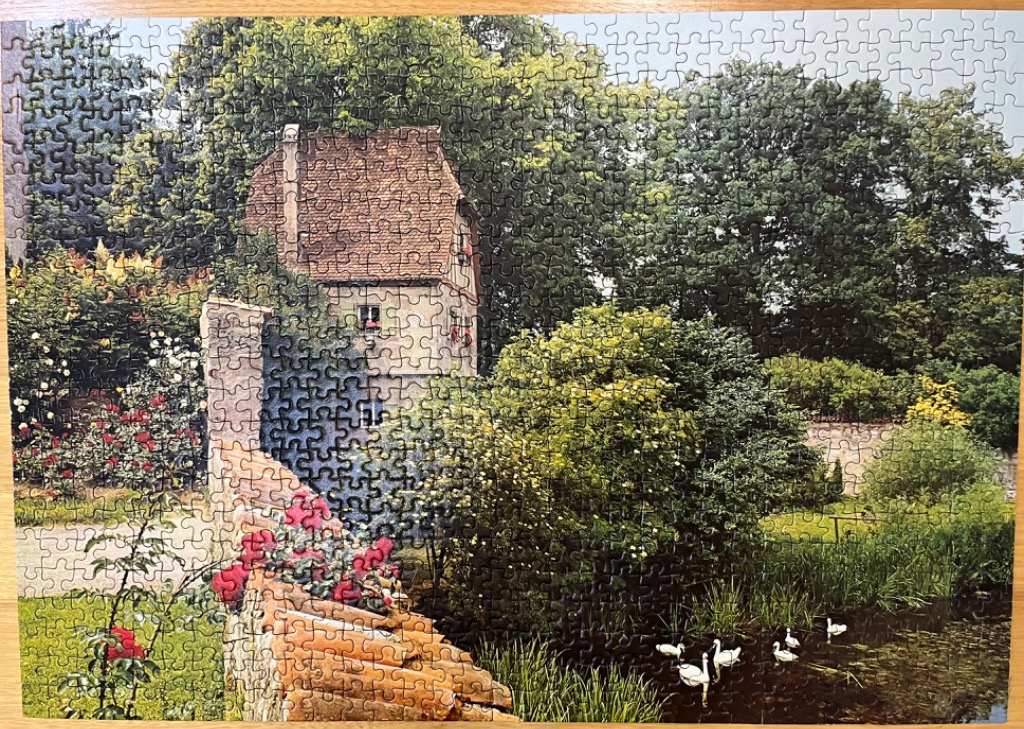 An old stone cottage with a tiled roof sits beside a stream and lush garden filled with flowers and trees.