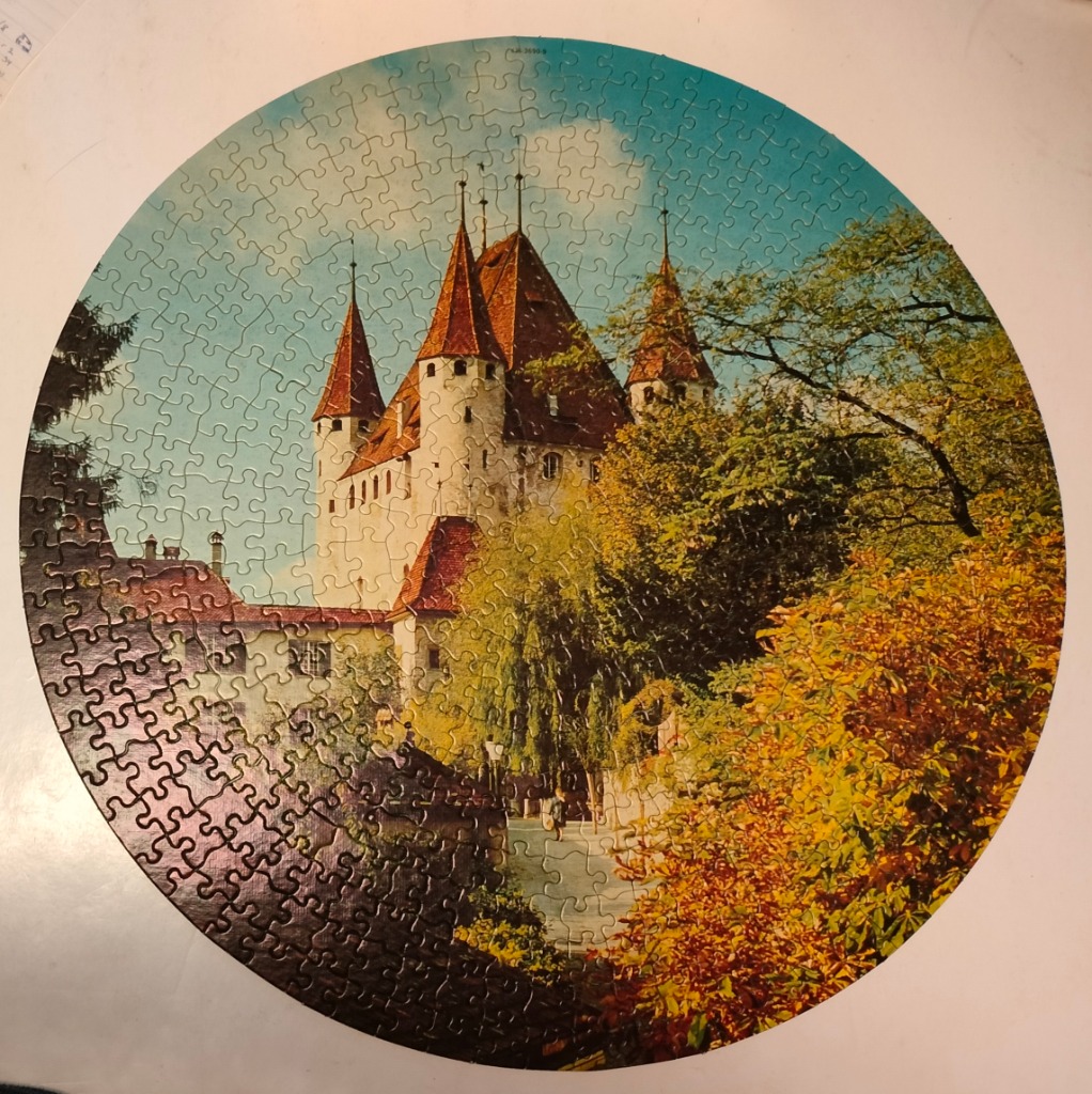 A circular image of a European-style stone castle with red roofs surrounded by lush green and autumn trees.