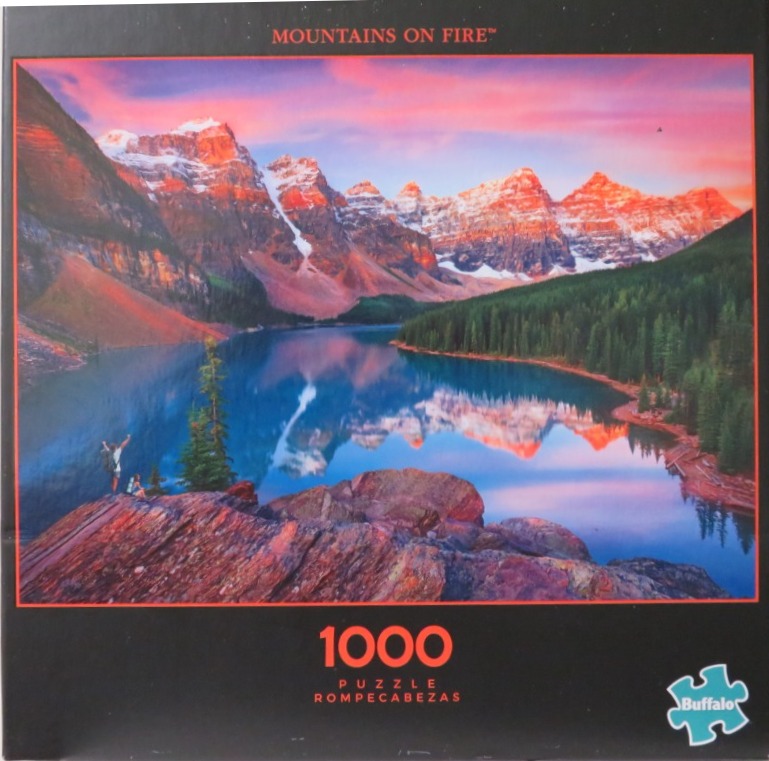 A hiker stands on a rocky outcrop looking over a deep blue alpine lake reflecting snowy mountains at sunset.