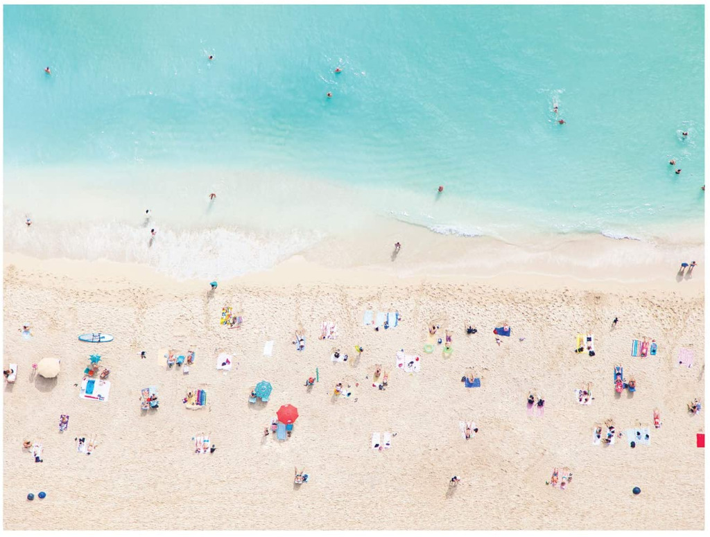 Aerial view of a sunny beach with swimmers in turquoise water and people relaxing on light sand.