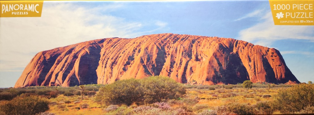 A panoramic landscape photograph of Uluru glowing in the bright Australian outback sun under a clear blue sky.