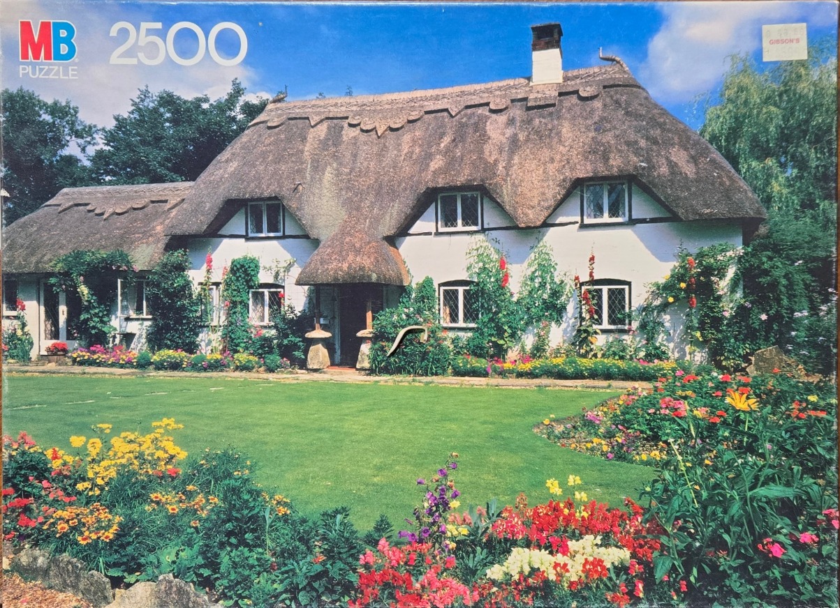 A charming white English cottage with a thatched roof, surrounded by a vibrant flower garden and a lush green lawn.