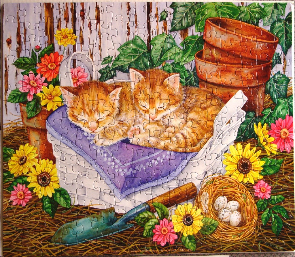 Two ginger kittens sleeping in a white basket surrounded by sunflowers and garden tools.