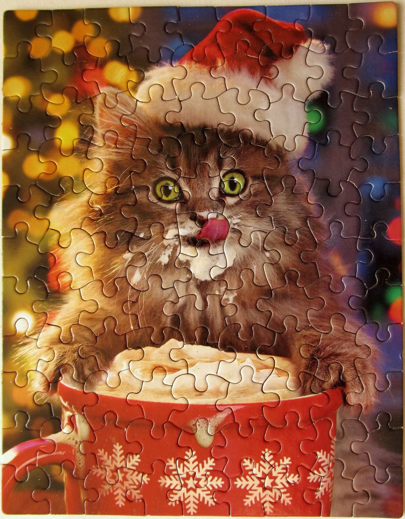 A fluffy cat wearing a Santa hat, licking its nose and peering out from a red mug filled with cocoa.