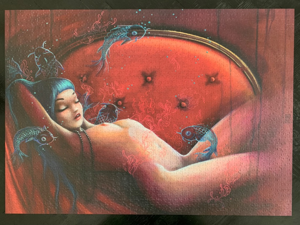 A woman with blue hair reclining on a tufted red velvet chair surrounded by floating, ethereal blue fish.
