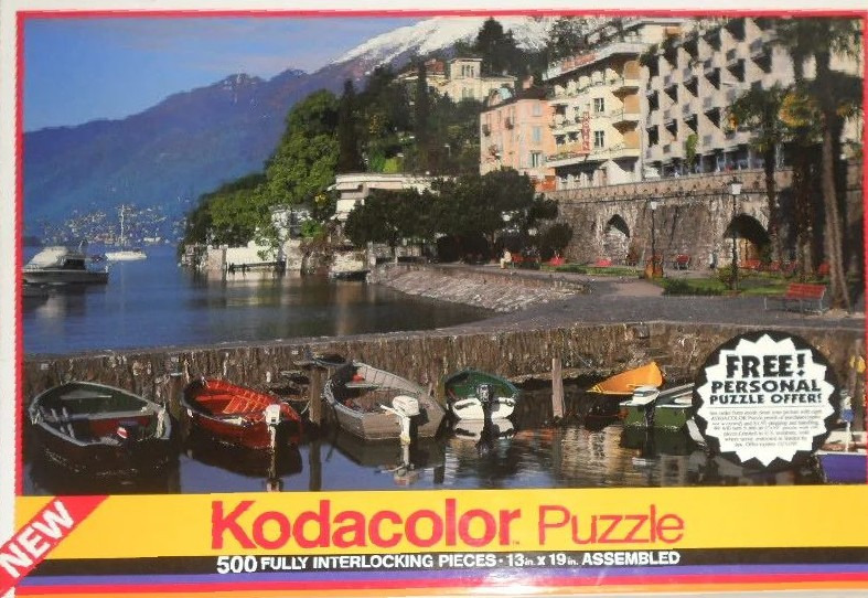 Lakeside scene with anchored rowboats and a hillside European resort town under snowy mountains.