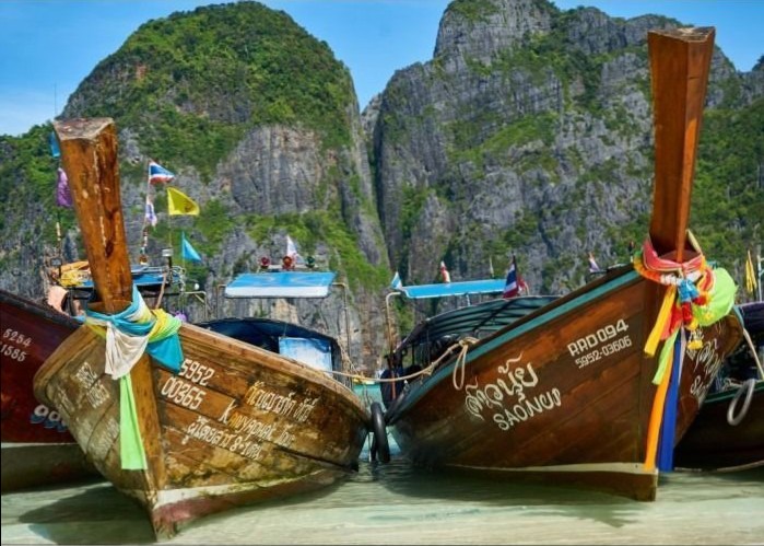 Two traditional wooden longtail boats docked on a beach against a backdrop of limestone cliffs.