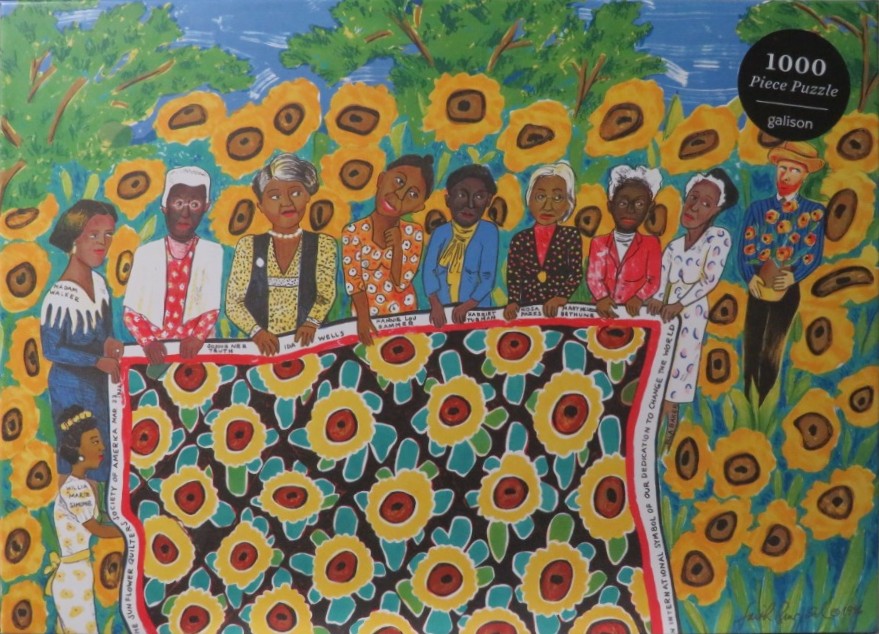 A group of influential Black women and a man holding a sunflower quilt, standing in a field of yellow sunflowers.