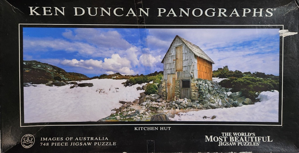 A panoramic landscape photograph of a weathered wooden hut standing in a snow-covered mountain clearing.