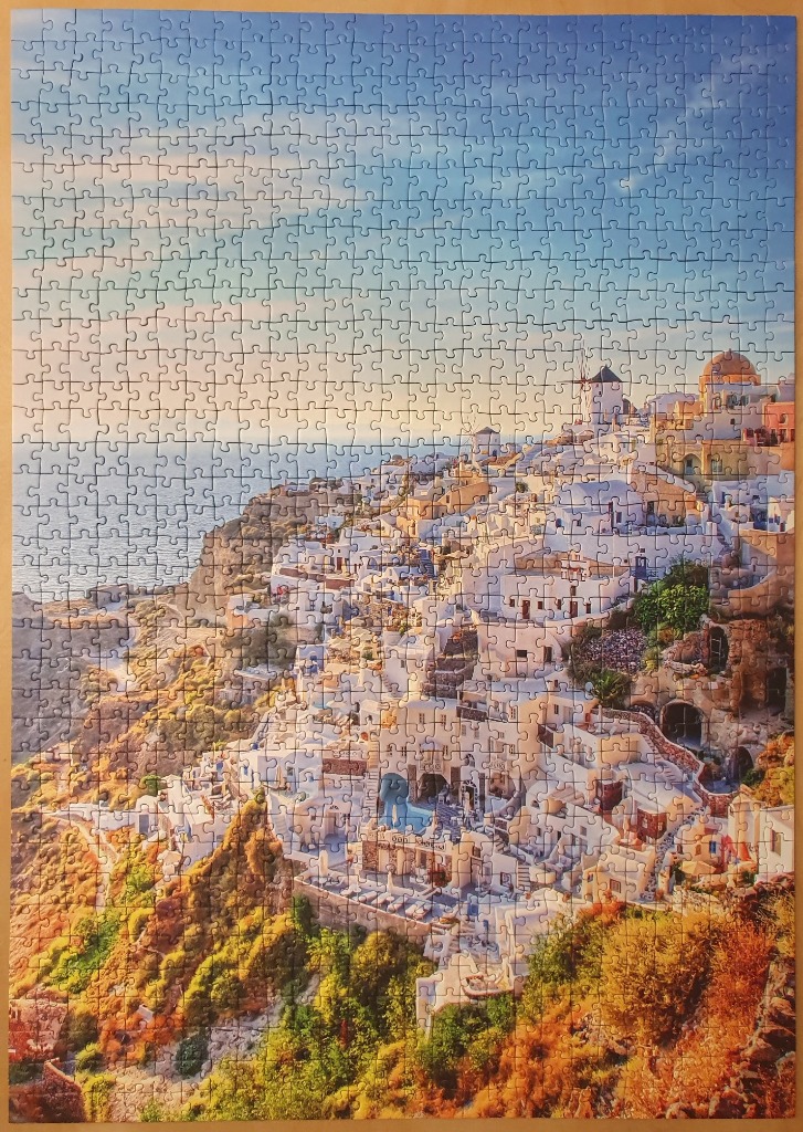 A scenic photograph of white buildings clinging to a cliffside in Santorini overlooking the ocean at sunset.