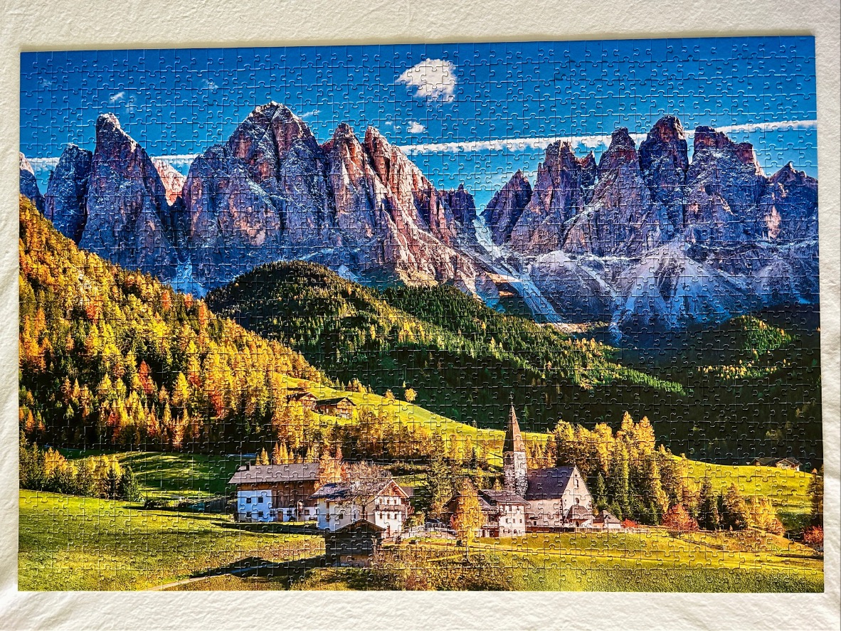 A panoramic landscape of the Dolomites mountain range rising above a small, idyllic alpine village in autumn.