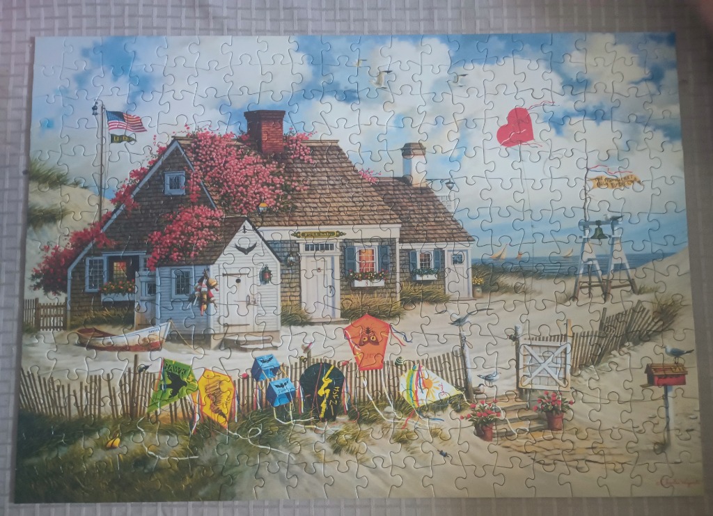 A coastal cottage surrounded by sand dunes, kites, and a flowering vine under a blue, cloudy sky.