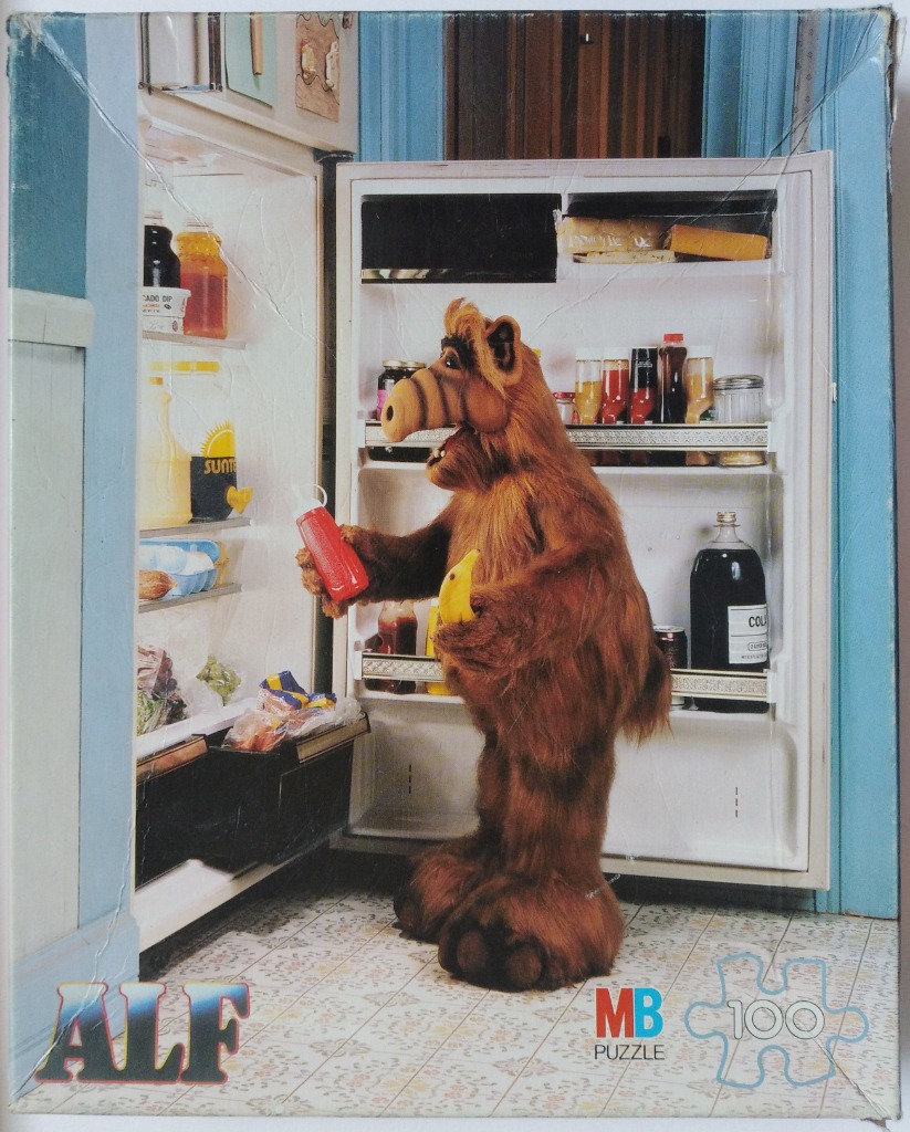 ALF the alien puppet standing in front of an open refrigerator holding a banana and a squeeze bottle of ketchup.
