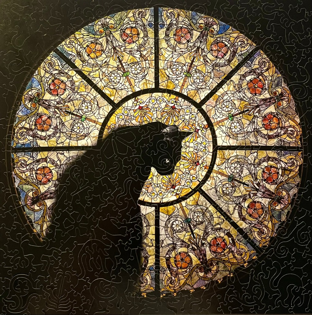 A silhouette of a black cat positioned in front of a colorful, ornate, circular stained glass window.