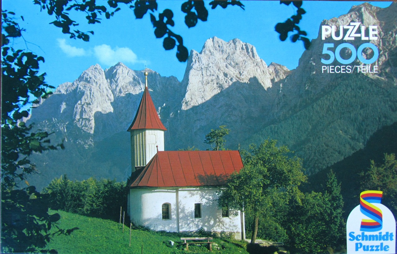A small white church with a red roof sits on a green hillside before a backdrop of towering, jagged alpine mountains.