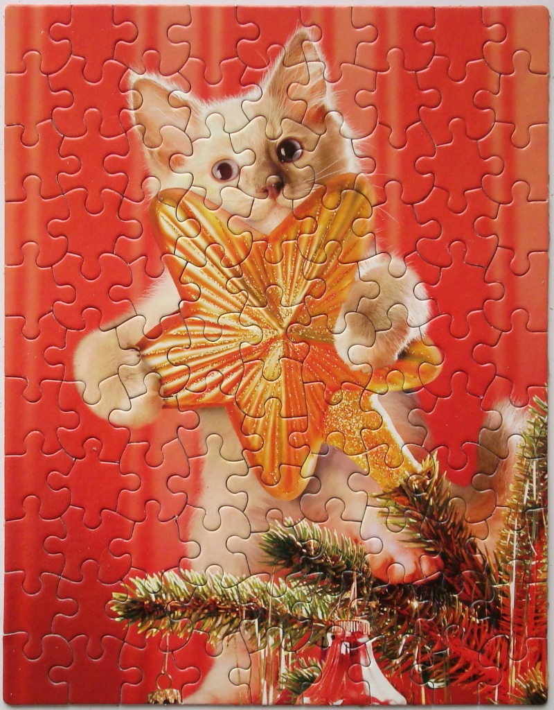 A fluffy white kitten holding a large, golden, star-shaped ornament in front of a bright red Christmas background.