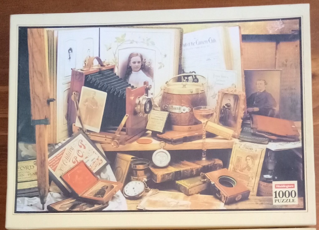 A still life featuring a vintage bellows camera, old photographs, antique books, and a wooden tobacco jar.
