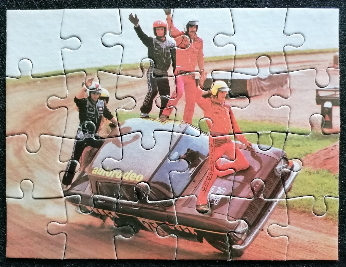 Four performers in racing jumpsuits stand on top of a car on a dirt track.