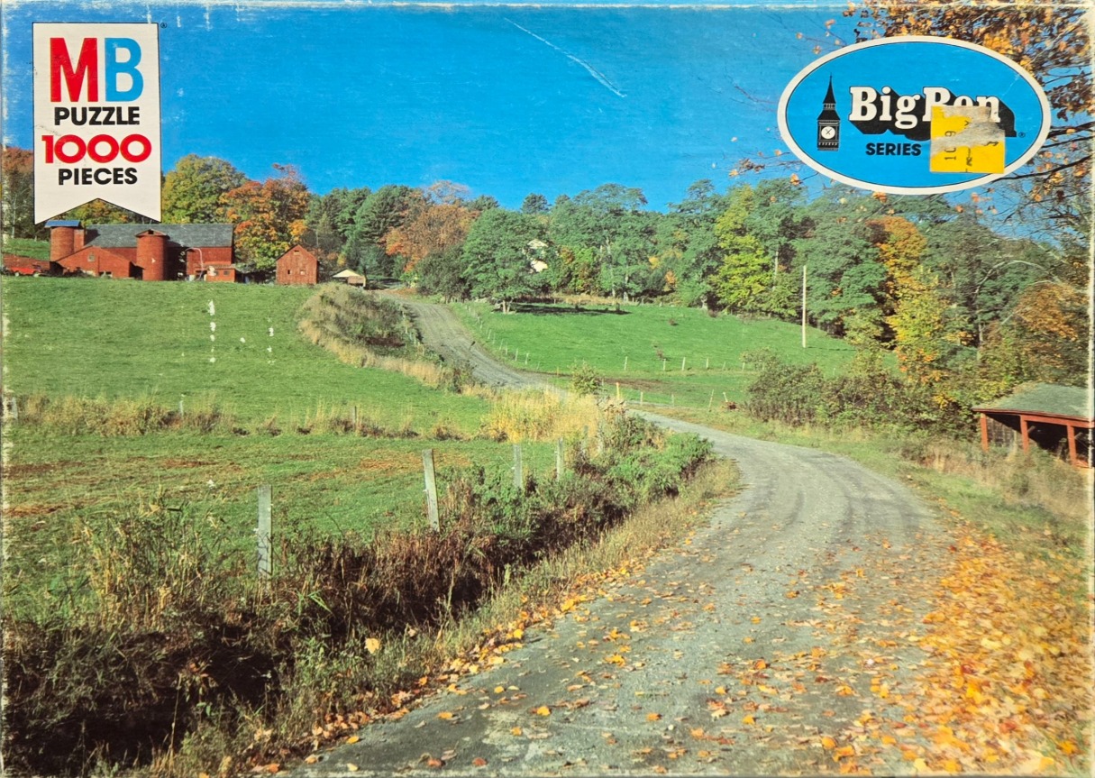 A rural landscape photograph of a dirt road winding toward a red barn and farmhouse on a sunny autumn day.